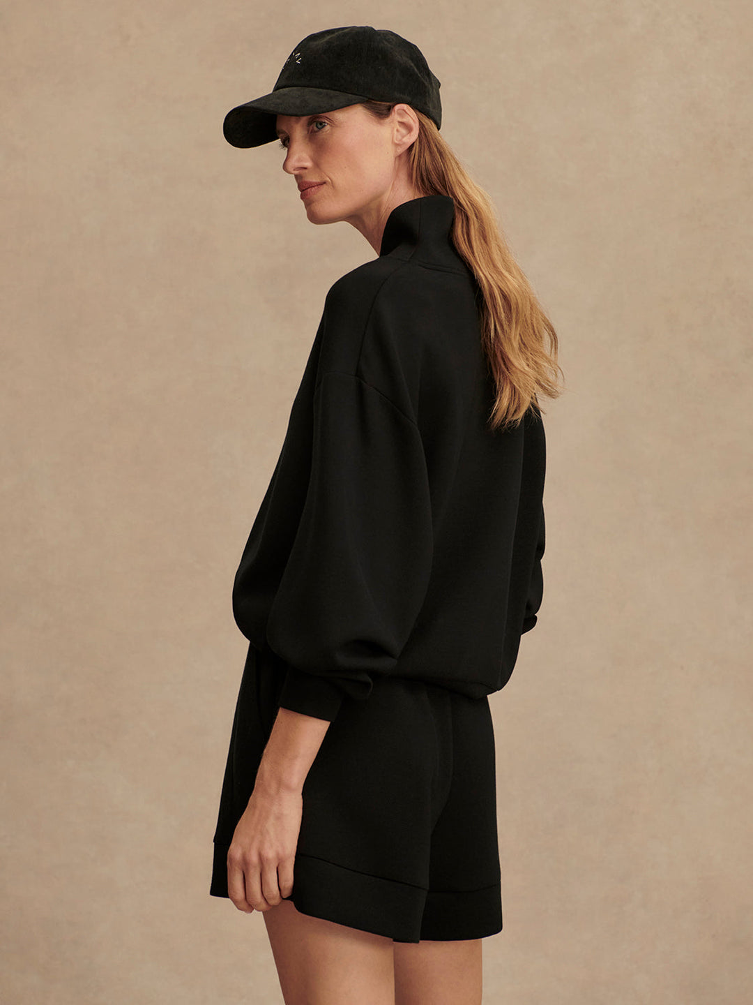 A woman in a black cap, black high-neck sweater with balloon sleeves, and Zertz ATRIUM HIGH RISE SHORT 4.5" INSEAM BLACK stands sideways against a beige background, looking ahead with her long hair down her back.