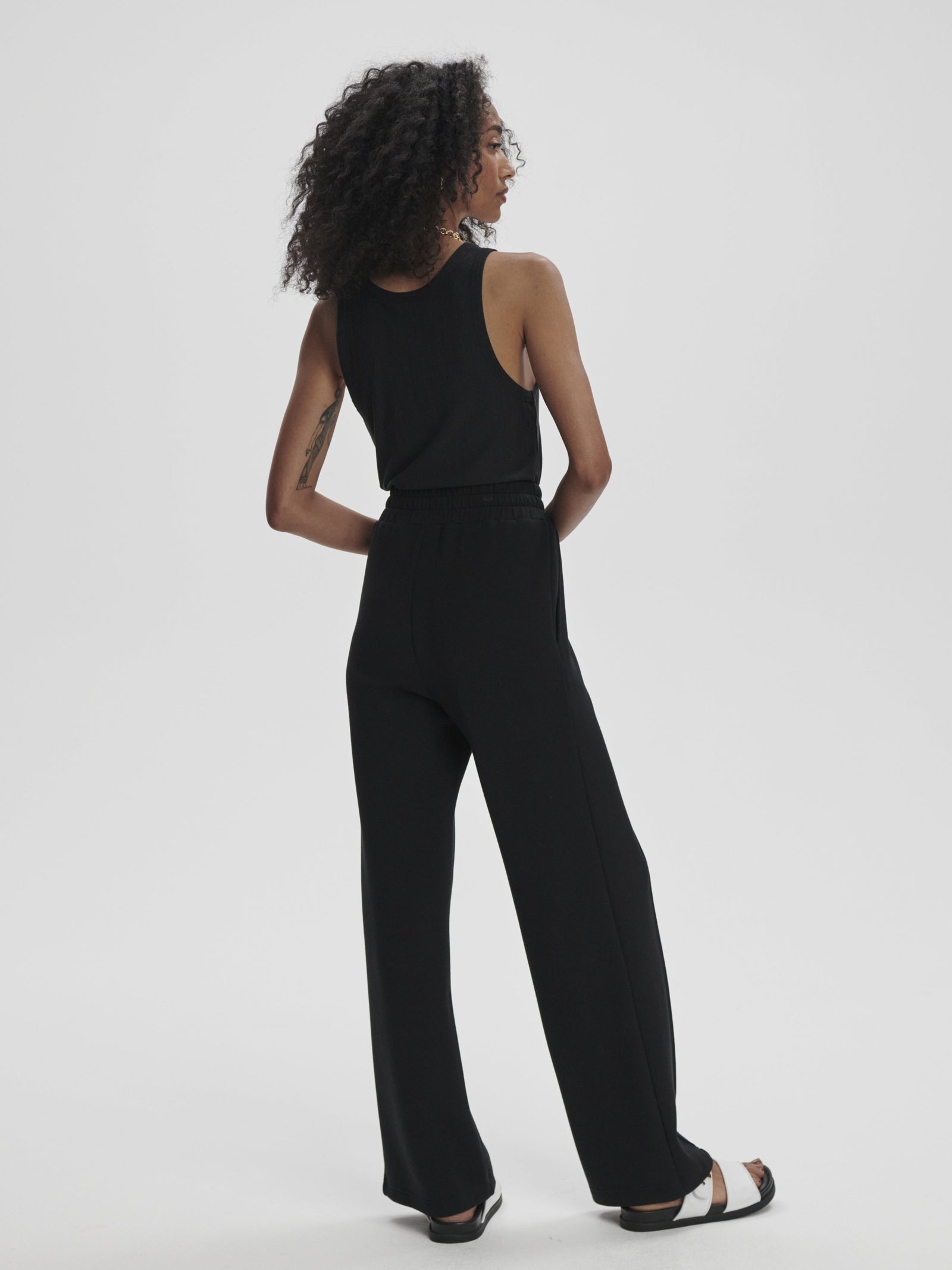 A woman with curly hair and a sleeveless black top wears Zertz THE WIDE LEG PANT BLACK and black sandals, standing facing away with hands on her lower back against a plain white background.