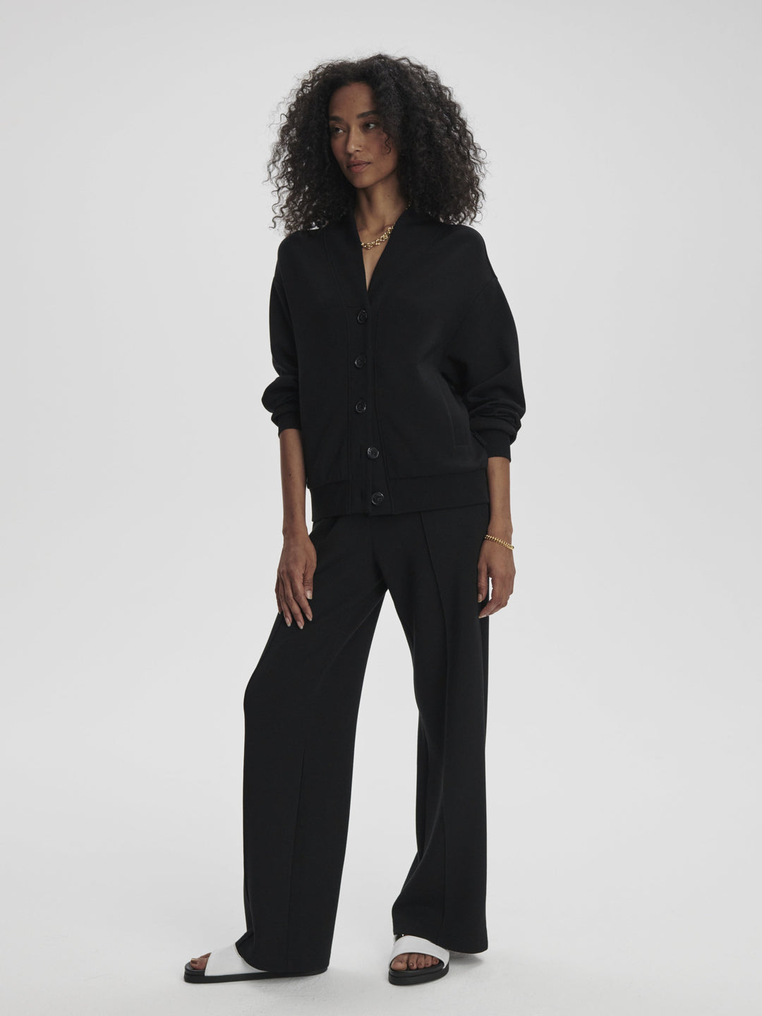 A woman with curly hair stands against a plain background, wearing a black button-up jacket, THE WIDE LEG PANT BLACK by Zertz, and black-and-white sandals. She has one hand in her pocket and wears minimal jewelry.