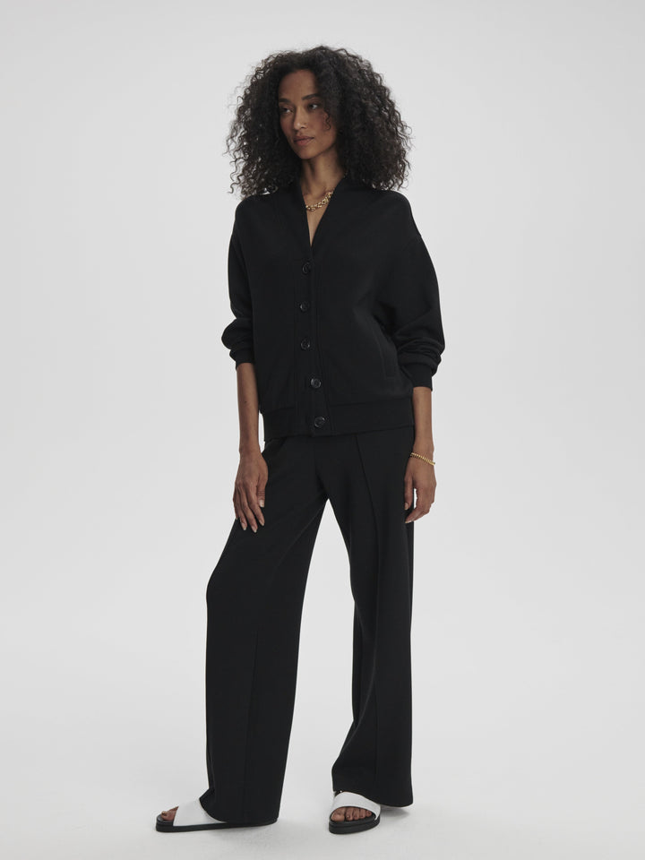 A woman with curly hair stands against a plain background, wearing a black button-up jacket, THE WIDE LEG PANT BLACK by Zertz, and black-and-white sandals. She has one hand in her pocket and wears minimal jewelry.