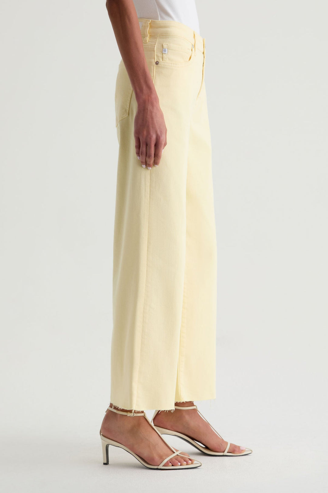 A person stands sideways in Zertz SAIGE WIDE LEG CROP jeans in pale yellow with a wide-leg fit and frayed hems, paired with white heeled sandals. The background is plain and light-colored.