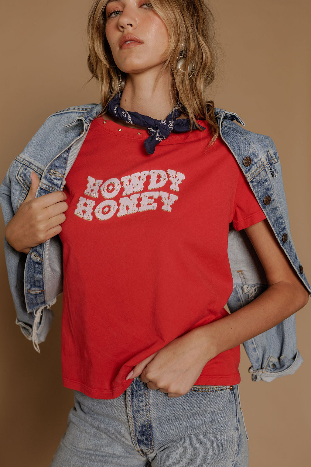 A woman wearing the Zertz HOWDY HONEY BOY TEE in red with light denim jeans, a denim jacket over her shoulders, dark blue bandana, and layered necklaces stands against a beige background.