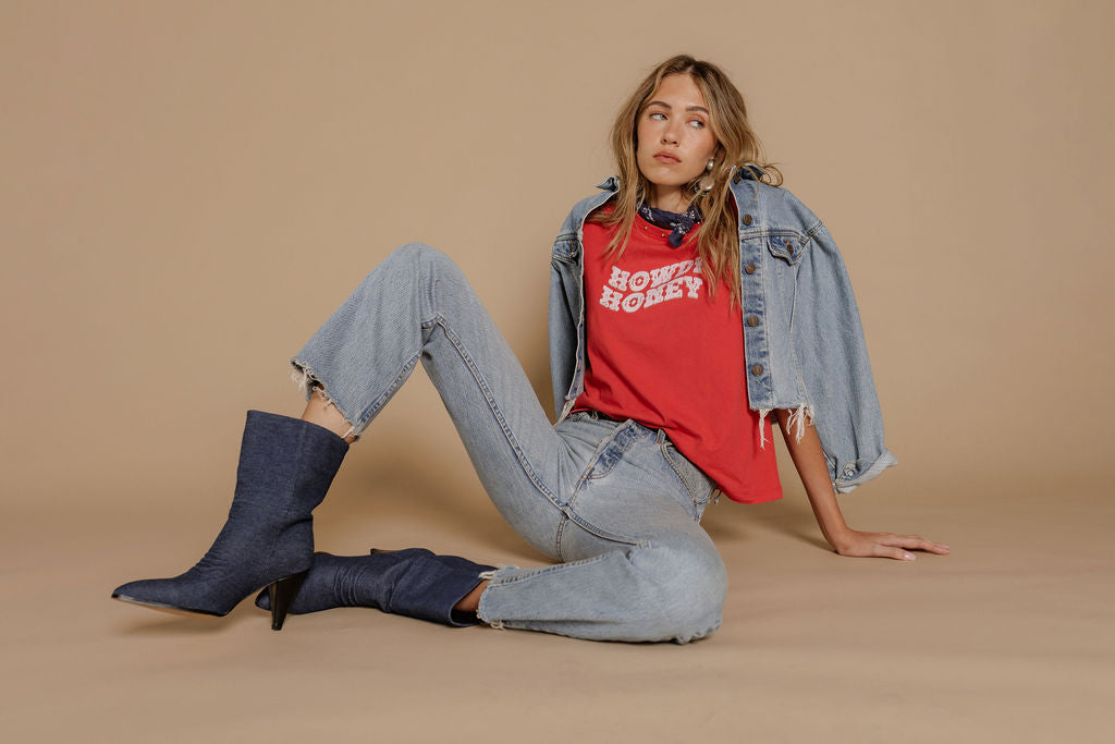 A woman with long, wavy hair sits on the floor against a beige background, wearing the Zertz HOWDY HONEY BOY TEE in red, light-wash jeans, a denim jacket over her shoulders, dark ankle boots, layered necklaces, and a patterned scarf.