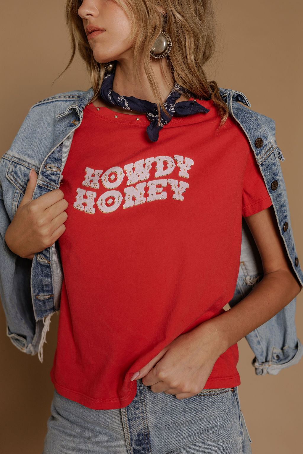 A woman wears the Zertz HOWDY HONEY BOY TEE in red, light denim jeans and a matching jacket over her shoulders, accessorized with a blue patterned bandana and delicate necklaces, posing against a beige background.