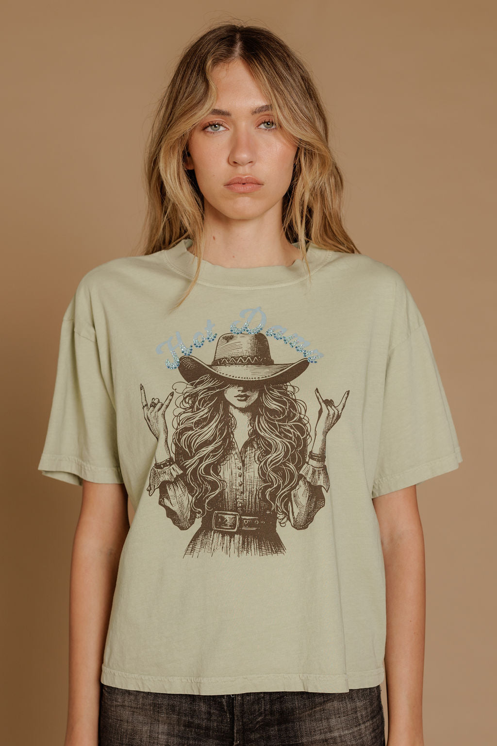 A woman with long wavy hair wears the Zertz HOT DAMN WIDE BOY TEE, a relaxed fit sage green graphic t-shirt featuring an illustrated woman in a hat and belted dress on a plain tan background.