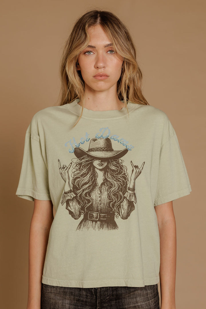A woman with long wavy hair wears the Zertz HOT DAMN WIDE BOY TEE, a relaxed fit sage green graphic t-shirt featuring an illustrated woman in a hat and belted dress on a plain tan background.