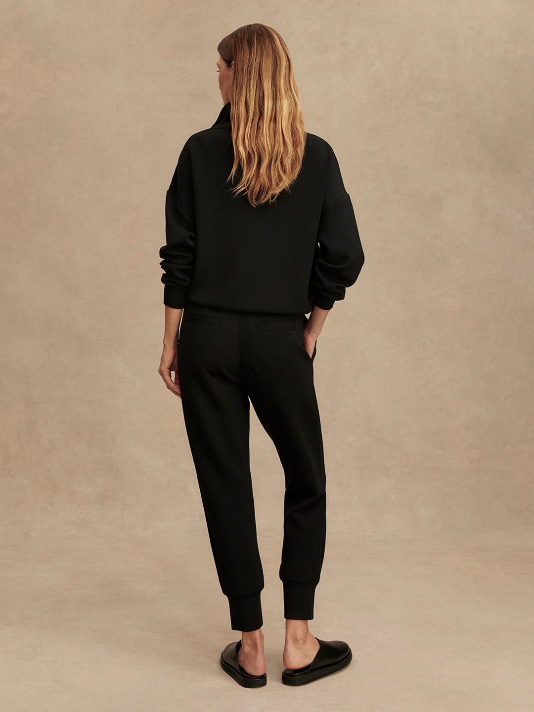 A woman with long, wavy blonde hair faces away from the camera, wearing a black sweatshirt and Zertz THE SLIM CUFF PANT 25-BLACK with tapered legs, paired with black slip-on shoes against a plain beige background.