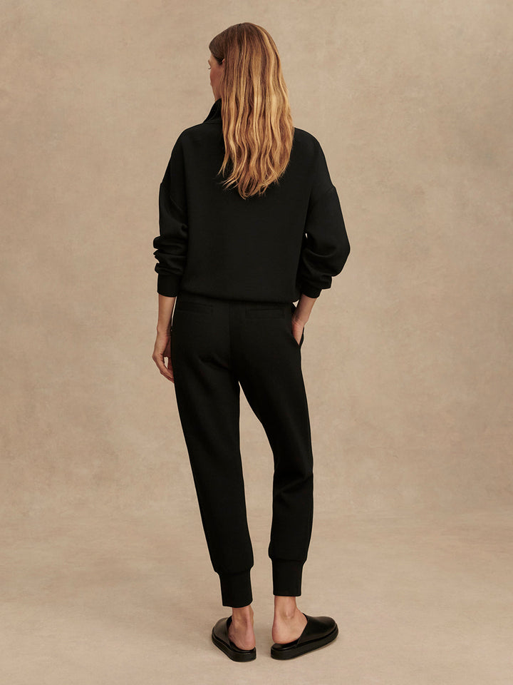 A woman with long, wavy blonde hair faces away from the camera, wearing a black sweatshirt and Zertz THE SLIM CUFF PANT 25-BLACK with tapered legs, paired with black slip-on shoes against a plain beige background.