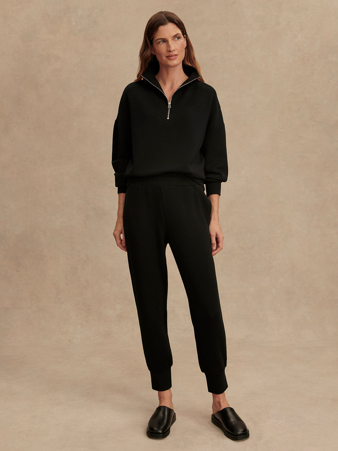 A woman with long brown hair stands against a beige background wearing Zertz's THE SLIM CUFF PANT 25-BLACK, a black zip-up sweater, and black slip-on shoes—her relaxed posture highlights this timeless wardrobe staple look.