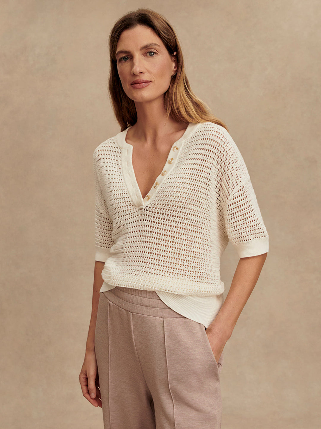 A woman with long brown hair wears the Zertz CALLIE KNIT TOP, made from breathable fabric, paired with light mauve pants, standing against a neutral beige background.