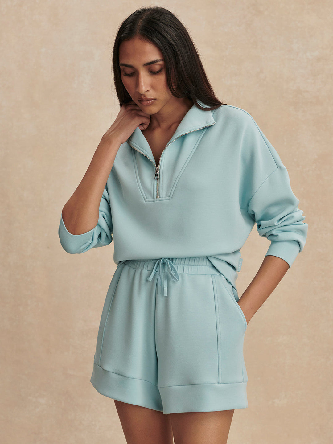 A woman wearing a light blue zip-up sweatshirt and Zertz ATRIUM HIGH RISE SHORT 4.5 INSEAM in Porcelain Blue stands against a beige background, looking down with one hand on her neck.