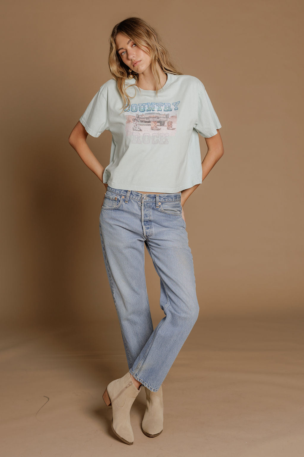 A woman stands against a tan backdrop wearing the Zertz COUNTRY ROCK WIDE BOY TEE in light blue, paired with light blue jeans and beige ankle boots, her hands in her pockets and head slightly tilted.