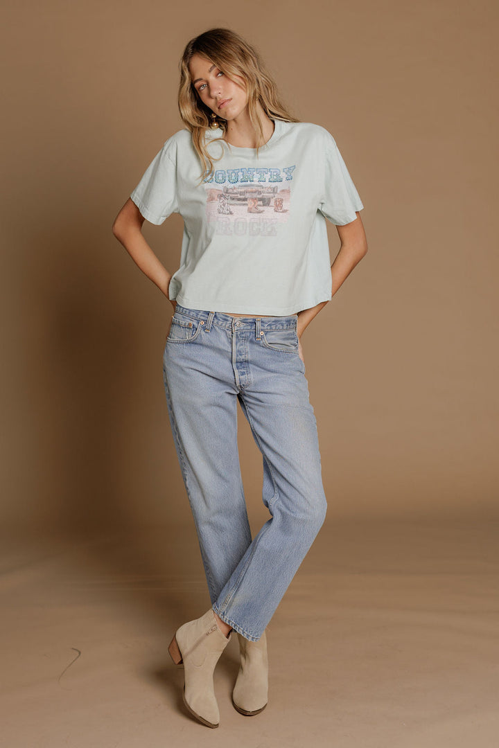 A woman stands against a tan backdrop wearing the Zertz COUNTRY ROCK WIDE BOY TEE in light blue, paired with light blue jeans and beige ankle boots, her hands in her pockets and head slightly tilted.