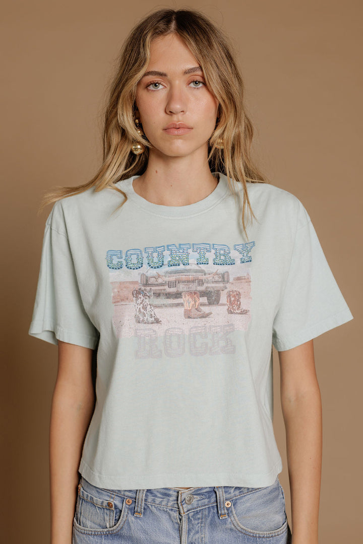 A woman with wavy blonde hair wears the Zertz COUNTRY ROCK WIDE BOY TEE, a premium cotton blend top with dog and truck graphics, along with blue jeans, standing against a plain brown backdrop.