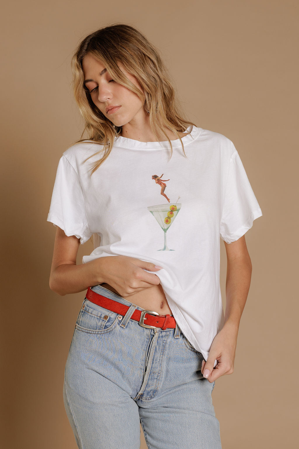 A woman wears the Zertz DIP INTO MARTINI TEE with a lounging graphic, paired with light blue jeans and a red belt, standing against a beige background—a fun choice for any cocktail lover’s casual style.