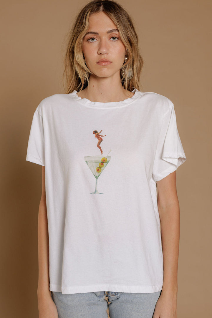 A woman with long, wavy blonde hair wears the Zertz DIP INTO MARTINI TEE—a white shirt featuring a martini glass with a small illustrated woman on the rim—standing against a plain beige background. Perfect for cocktail lovers.