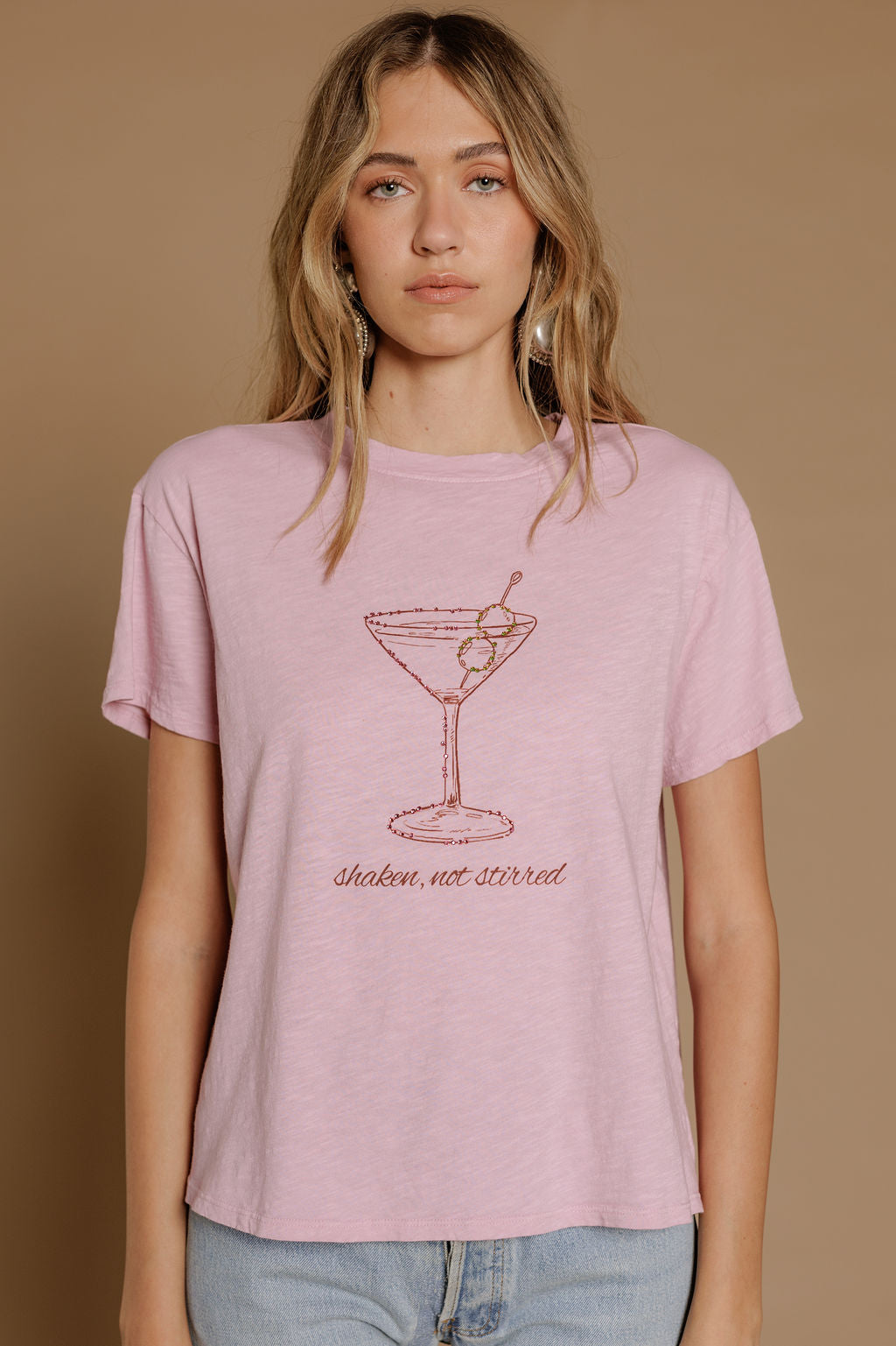 A woman with wavy blonde hair wears the Zertz SHAKEN NOT STIRRED SLUB CAP TEE in light pink, featuring a martini glass and "shaken, not stirred" text, standing against a plain beige background.