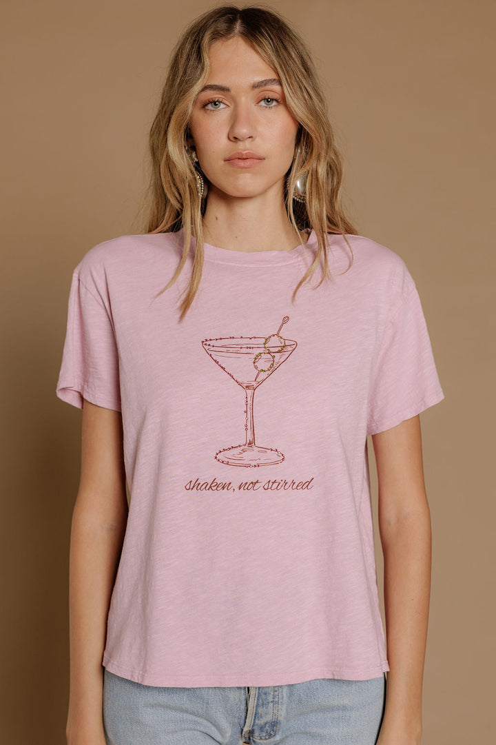 A woman with wavy blonde hair wears the Zertz SHAKEN NOT STIRRED SLUB CAP TEE in light pink, featuring a martini glass and "shaken, not stirred" text, standing against a plain beige background.