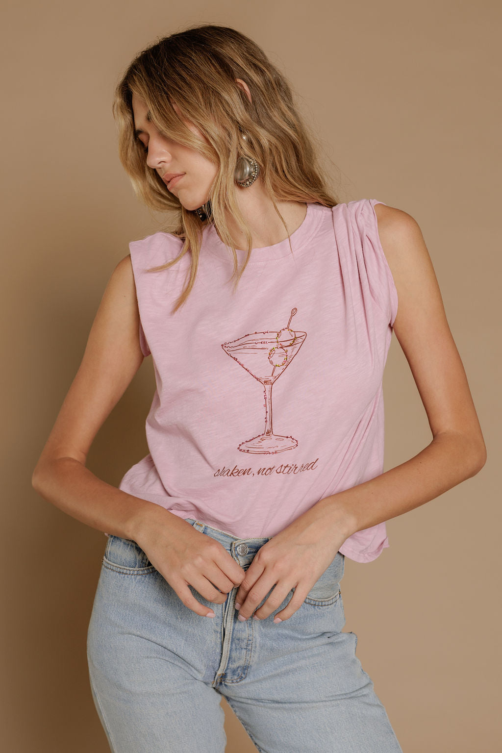 A woman models the Zertz SHAKEN NOT STIRRED SLUB CAP TEE featuring a martini glass graphic and "shaken, not stirred" text, paired with light blue jeans against a beige background, looking down to her left.