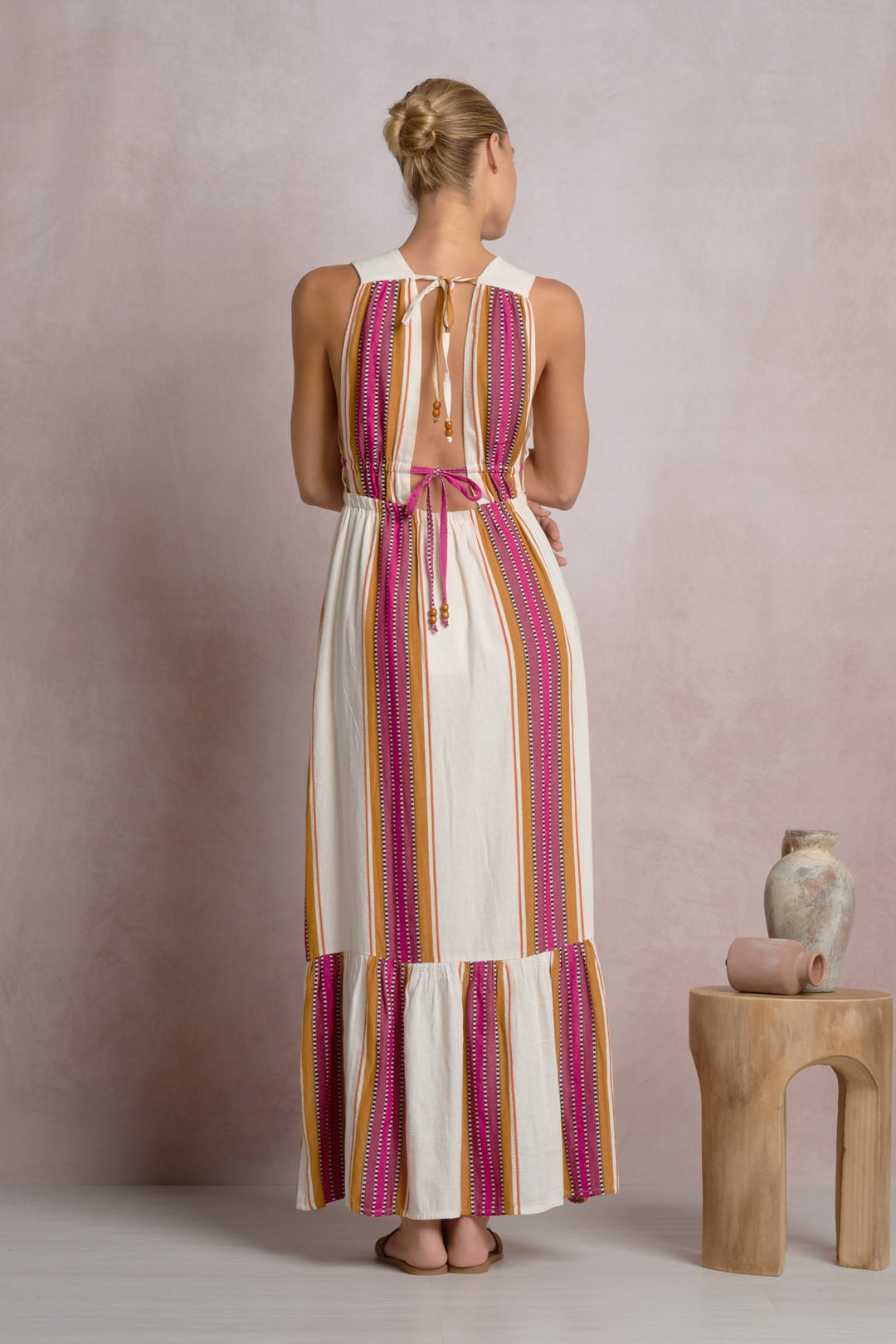 A woman faces away, wearing the Zertz RUFFLE MAXI HALTER DRESS—a sleeveless, ankle-length dress with pink, orange, and white vertical stripes, an open back with ties. She&