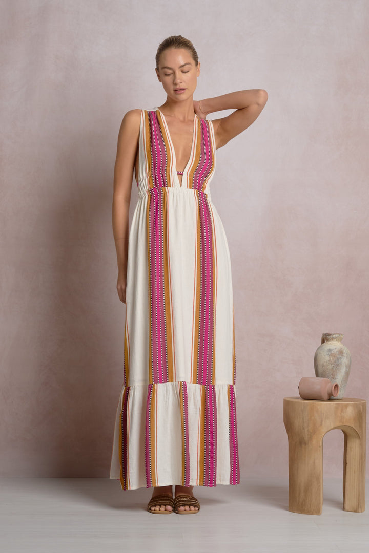 A woman poses near a rustic vase and stool, wearing the Zertz RUFFLE MAXI HALTER DRESS—sleeveless with a deep V-neck, ruffled hem, and long white maxi silhouette featuring vertical pink, orange, and yellow stripes.