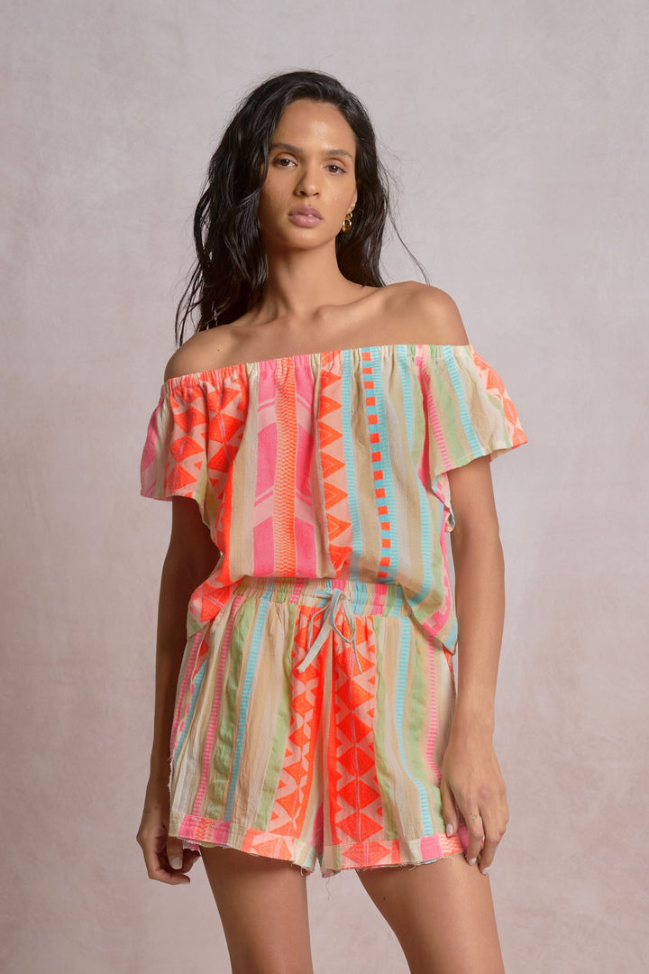 A woman with long dark hair models the Zertz OFF SHOULDER RUFFLE SLEEVE TOP with colorful geometric patterns, perfect for festivals, as she stands against a light beige background.