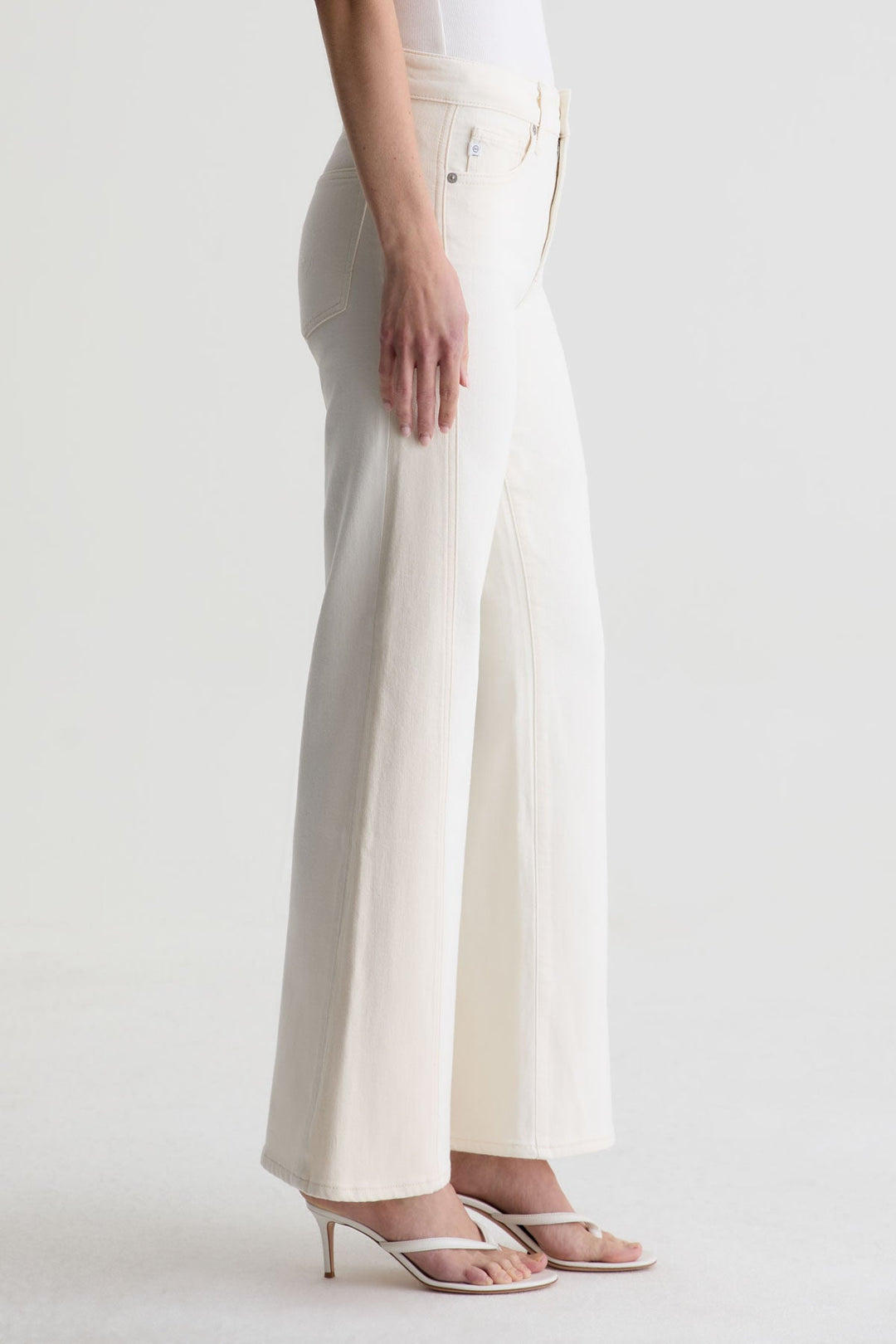 A person shown from the side wears Zertz’s SAIGE WIDE LEG MID RISE jeans and white heeled sandals, with only the lower half of their body visible against a plain light background.