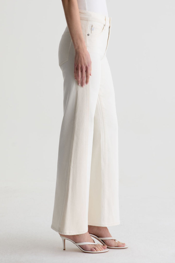 A person shown from the side wears Zertz’s SAIGE WIDE LEG MID RISE jeans and white heeled sandals, with only the lower half of their body visible against a plain light background.