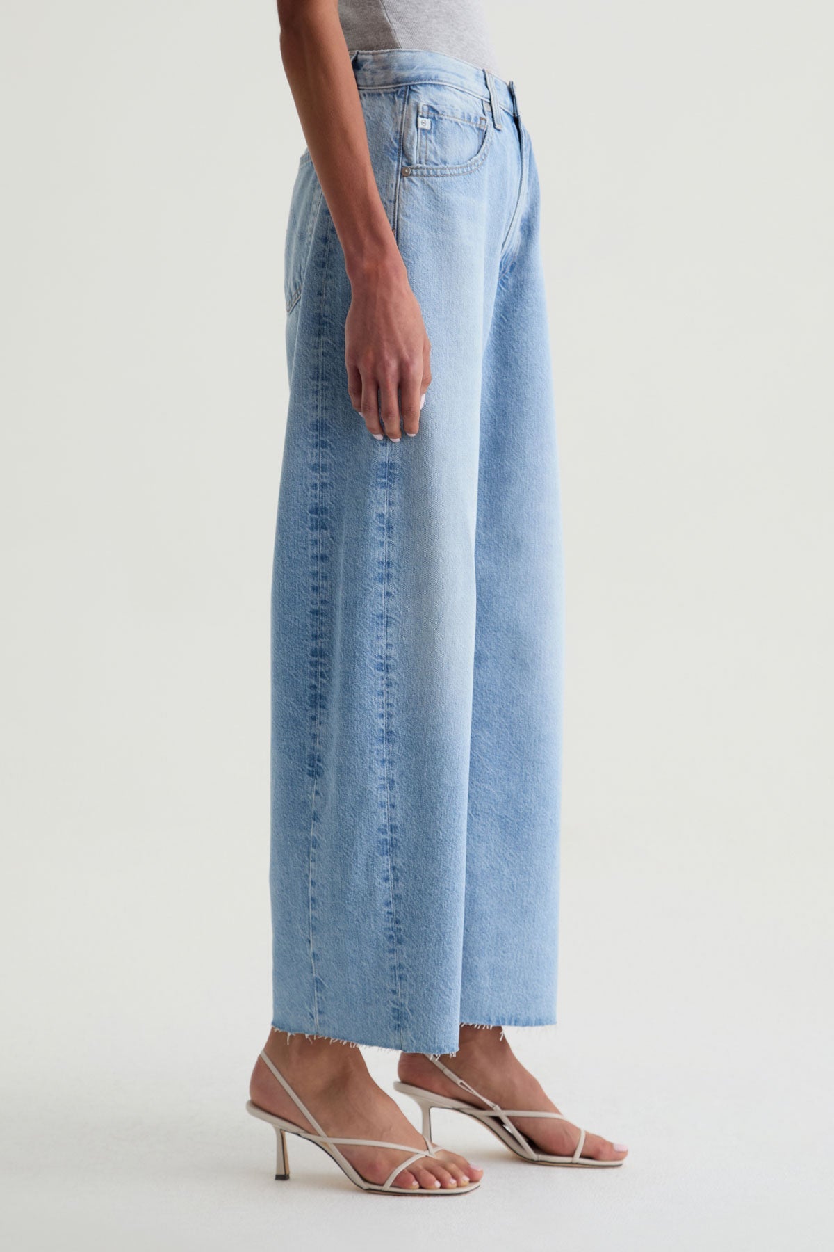 A person in Zertz HATTIE CROP BARREL JEAN GESTURE—light blue, relaxed fit with raw hem—paired with a gray top and white heeled sandals stands against a plain light background.