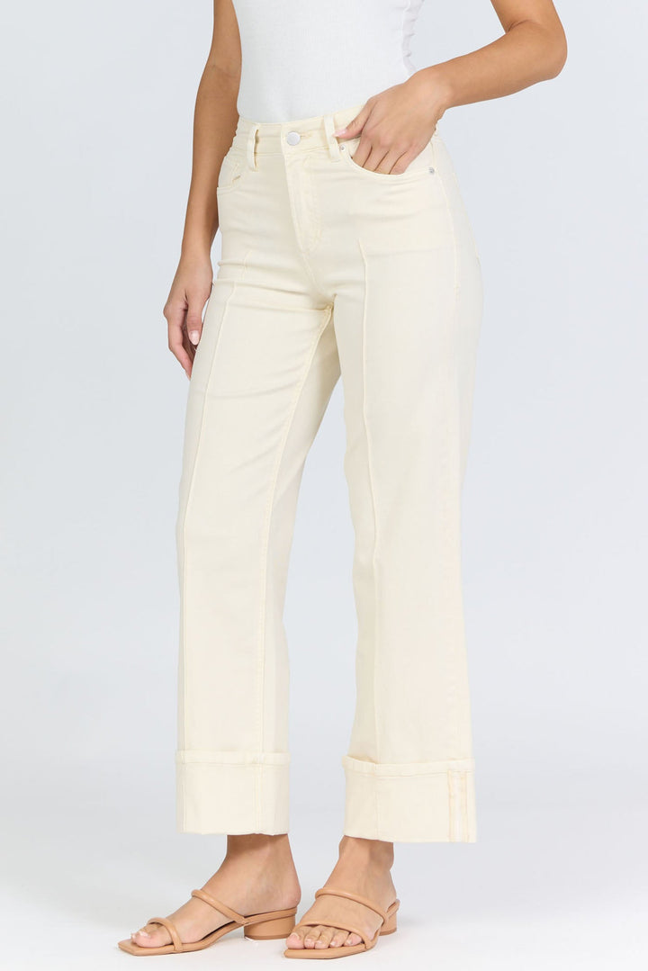 A woman in Zertz’s HOLLY JEAN high-waisted, off-white wide-leg jeans with cuffed hems, a white sleeveless top, and tan slide sandals radiates classic style against a simple light background.
