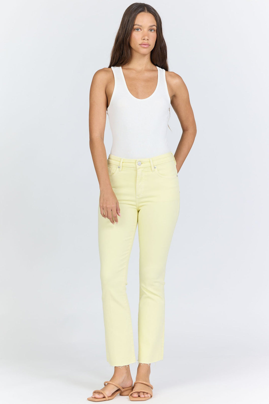 A woman with long brown hair wears a sleeveless white top, tan sandals, and Zertz JEANNE JEAN yellow high-waisted denim jeans in a comfortable fit, standing against a plain light background.