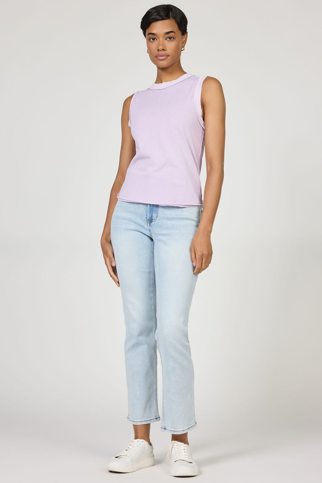 A woman with short hair faces forward against a plain background, wearing a sleeveless lavender Zertz ZO TANK made from breathable fabric, paired with light blue jeans and white sneakers.