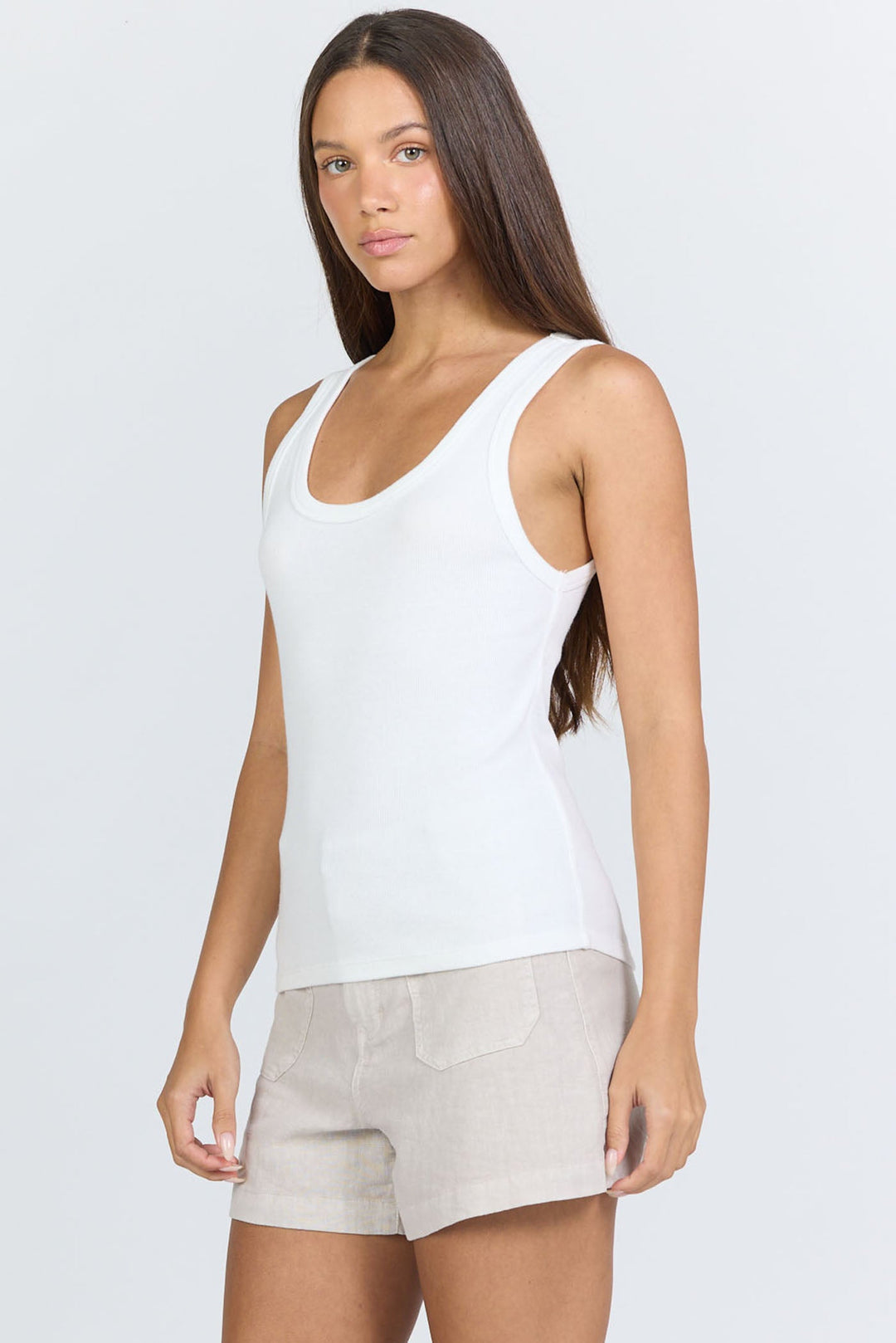 A woman with long brown hair wears the Zertz LOUISE TANK and beige shorts, standing against a light gray background with a neutral expression. This minimalist look highlights the versatility of this essential wardrobe staple.