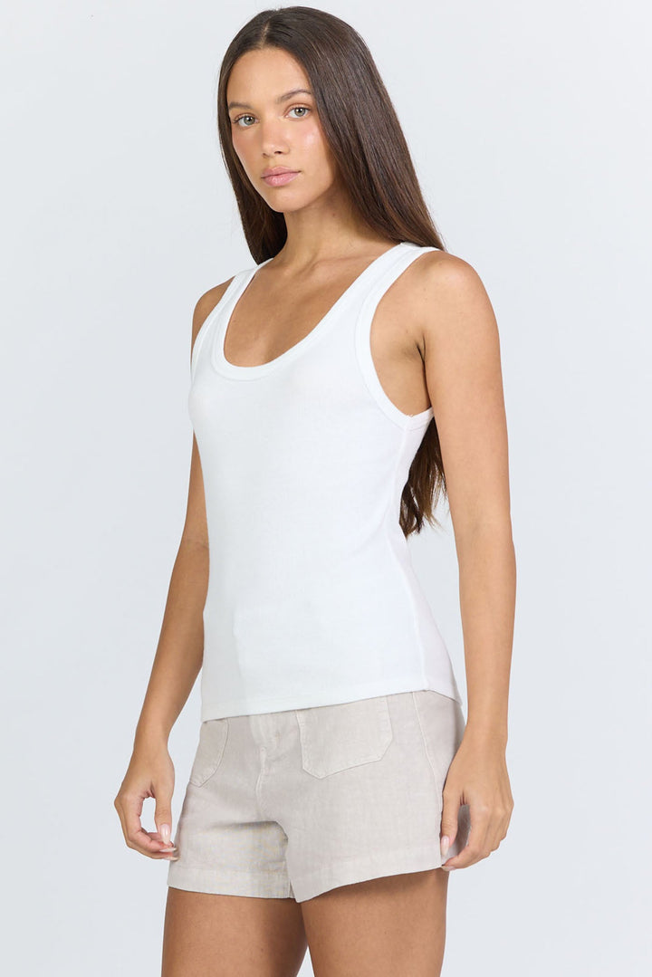A woman with long brown hair wears the Zertz LOUISE TANK and beige shorts, standing against a light gray background with a neutral expression. This minimalist look highlights the versatility of this essential wardrobe staple.