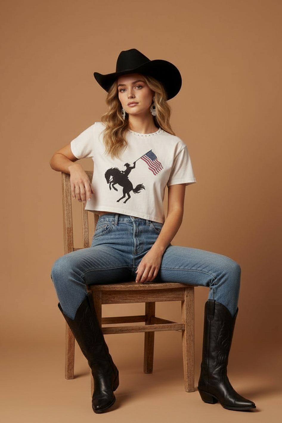 A woman in a black cowboy hat, blue jeans, and black boots sits on a wooden chair against a plain brown background, wearing the Zertz AMERICAN COWBOY WIDE BOY TEE with a rodeo and American flag graphic.
