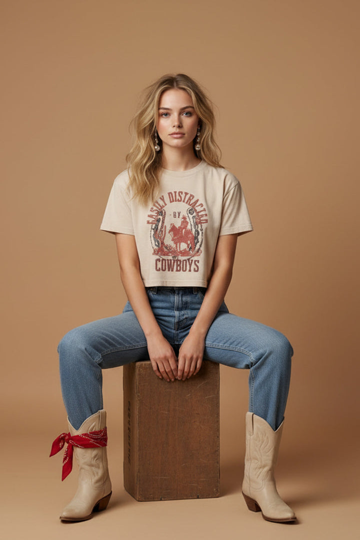 A woman with long blonde hair sits on a wooden box against a beige background, wearing the Zertz EASILY DISTRACTED WIDE BOY TEE in an oversized fit, blue jeans, cowboy boots, and a red bandana around her right ankle for casual comfort.