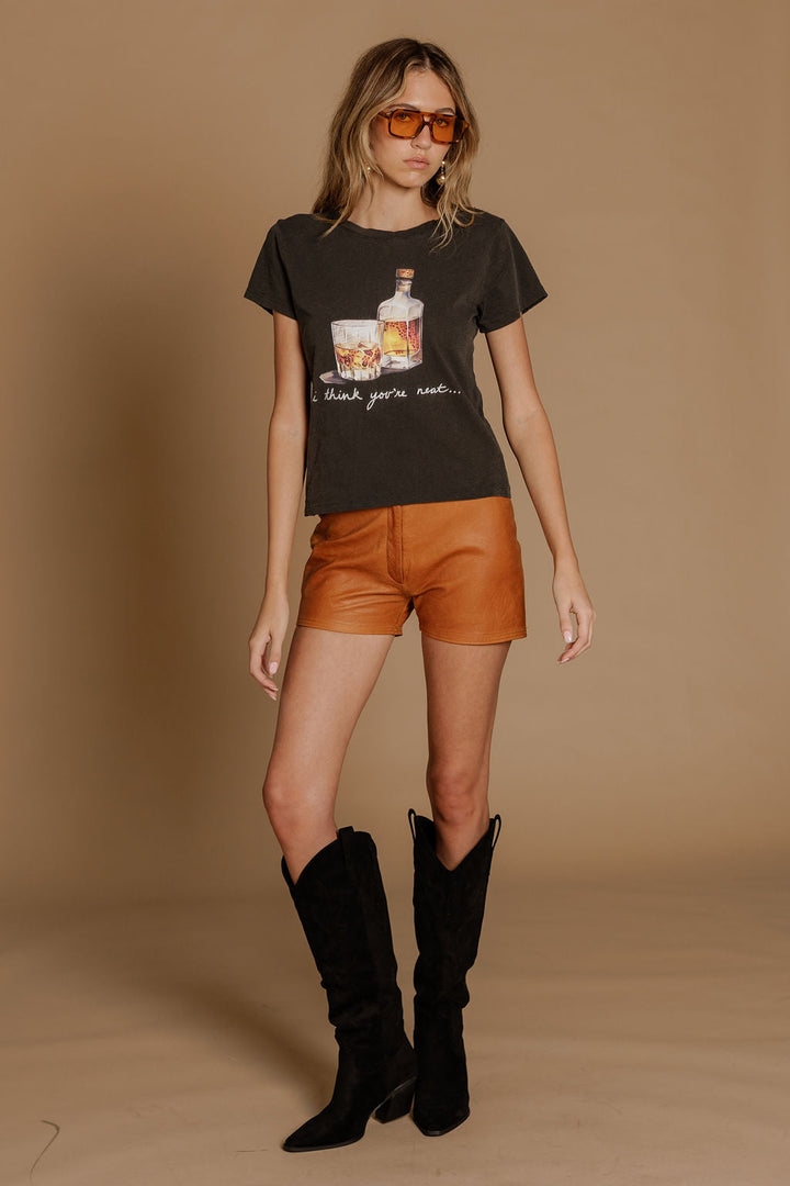 A woman in orange sunglasses, the Zertz I THINK YOUR NEAT SLIM TEE, brown leather shorts, and tall black boots stands against a plain beige background.