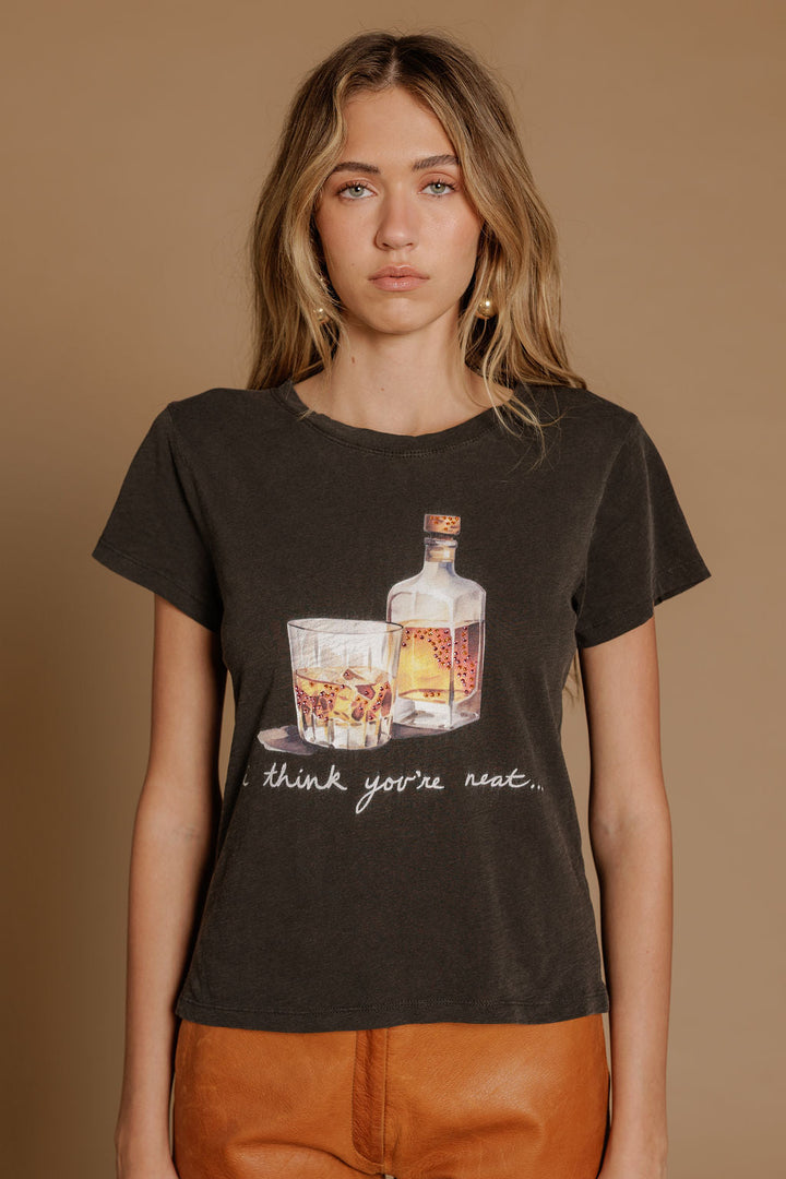 A woman with wavy blonde hair models the Zertz I THINK YOUR NEAT SLIM TEE, showcasing whiskey bottle and glasses illustrations. She stands against a tan background, styled with brown pants.