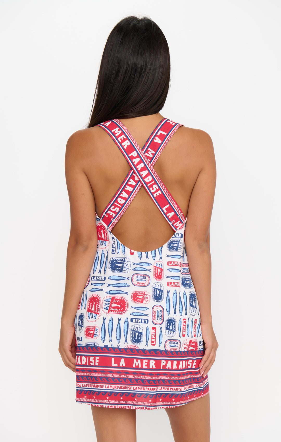 A woman faces away, wearing the Zertz DETOUR MINI DRESS—a sleeveless, versatile mini dress with a low open back, crisscrossed straps, red, blue, and white patterns, and “LA MER PARADISE” on the straps and hem.