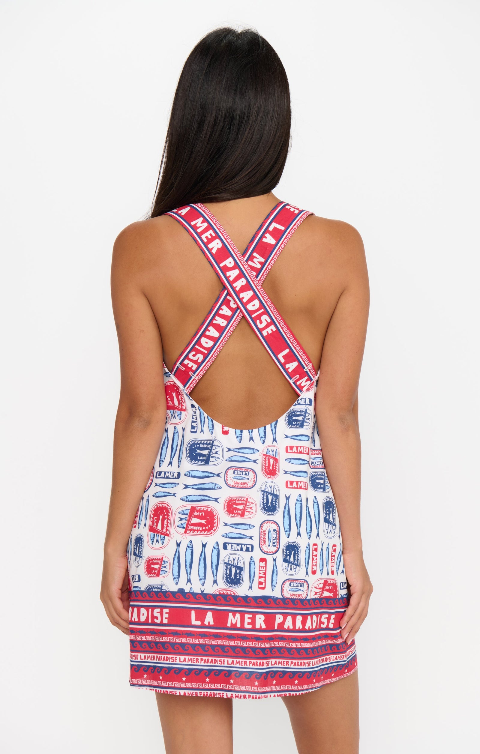 A woman faces away, wearing the Zertz DETOUR MINI DRESS—a sleeveless, versatile mini dress with a low open back, crisscrossed straps, red, blue, and white patterns, and “LA MER PARADISE” on the straps and hem.