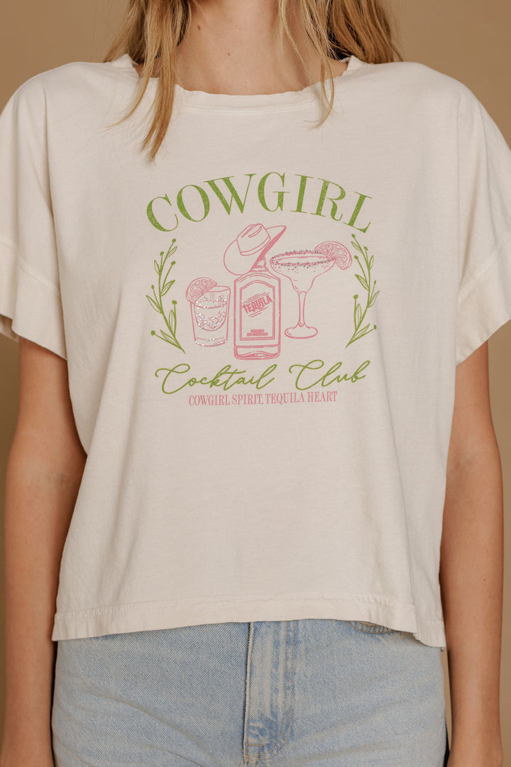 A woman wears the Zertz COWGIRL COCKTAIL HI LOW TEE, featuring “Cowgirl Cocktail Club” with a hat, tequila bottle, margarita glass, and lime graphic, paired with light blue high-waisted jeans.