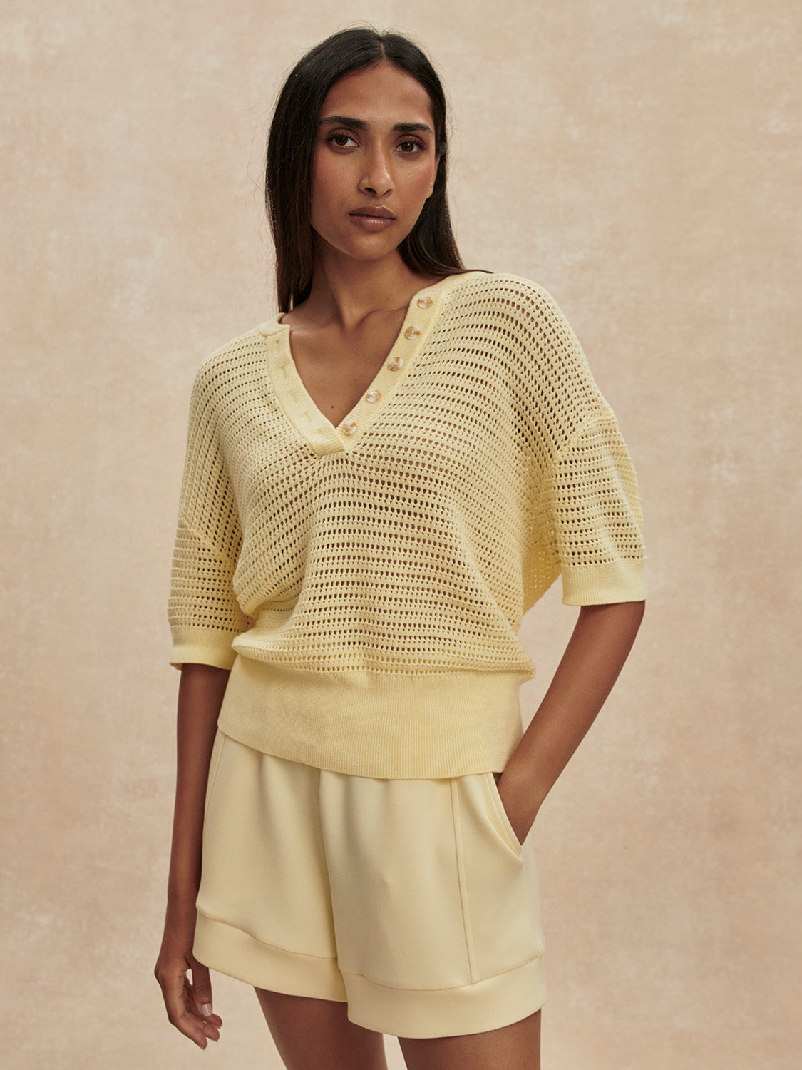 A woman with long dark hair wears the Zertz CALLIE KNIT TOP—a light yellow, airy V-neck knit with button details—paired with matching high-waisted shorts, standing against a beige textured background.
