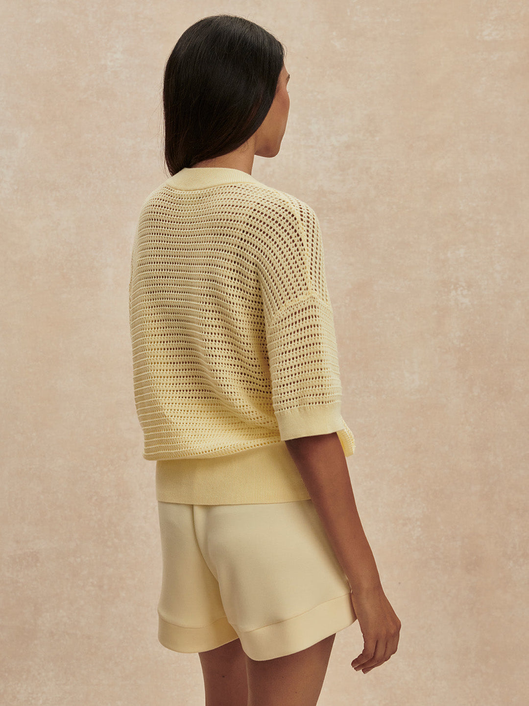 A woman with long dark hair, seen from behind, wears the Zertz CALLIE KNIT TOP—an effortlessly versatile pale yellow piece—paired with matching shorts against a neutral beige background.
