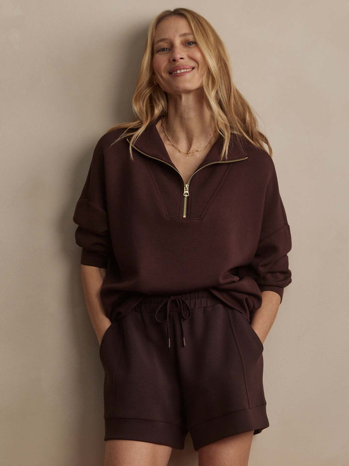 A woman with long blonde hair smiles in a dark brown zip-up and Zertz ATRIUM HIGH RISE SHORT 4.5, standing against a beige wall—the perfect look for any warm-weather wardrobe.