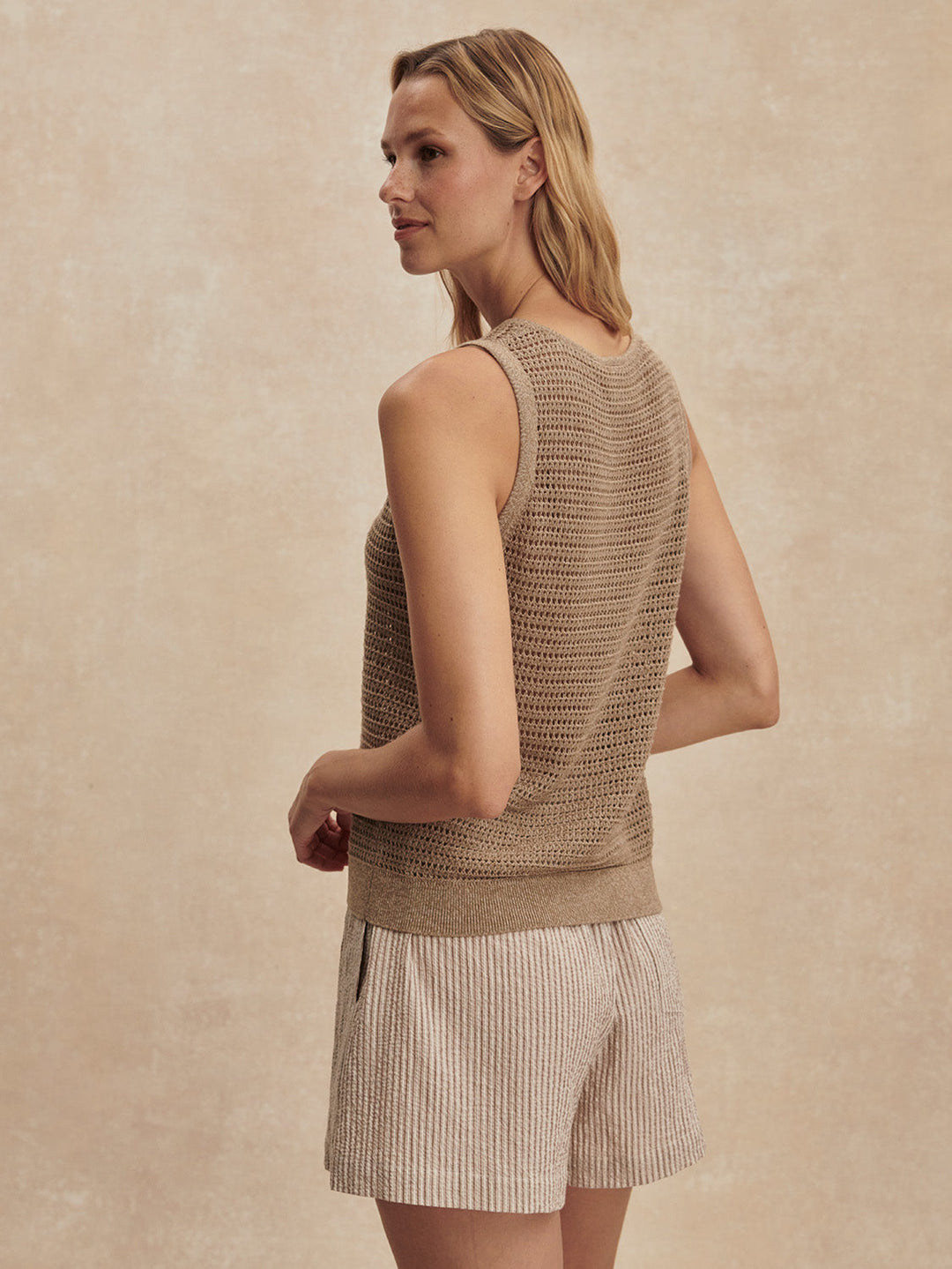A woman with light hair stands sideways, looking over her shoulder. She wears the Zertz RAMON KNIT TANK-SEPIA TWIST against a neutral, textured background for an effortlessly stylish look.