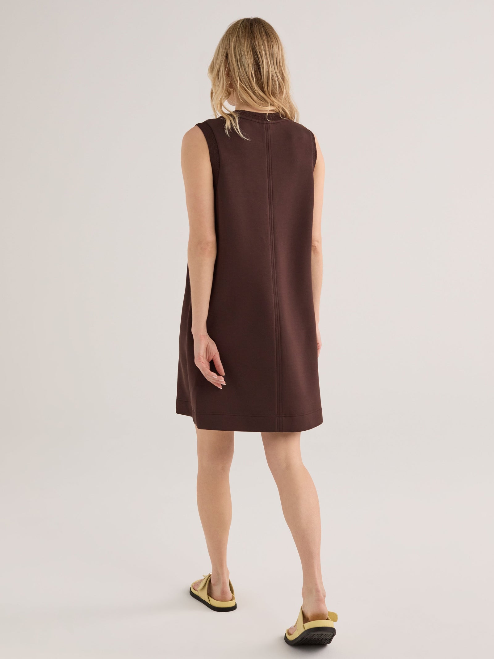 A woman with wavy blonde hair stands with her back to the camera, wearing the Zertz ELBA TANK MINI DRESS-COFFEE BEAN and flat sandals against a plain, light background.