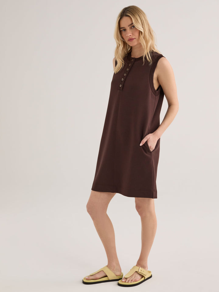 A woman wears the Zertz ELBA TANK MINI DRESS-COFFEE BEAN, a sleeveless dark brown mini dress with button details and pockets, styled with light yellow sandals, posing against a plain background.